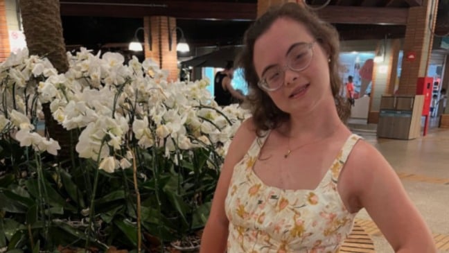 Jovem mulher com óculos e vestido floral posa sorridente ao lado de orquídeas brancas em ambiente interno bem iluminado.