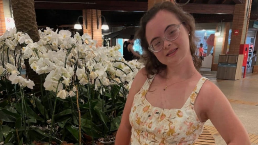 Jovem mulher com óculos e vestido floral posa sorridente ao lado de orquídeas brancas em ambiente interno bem iluminado. Jovem mulher com óculos e vestido floral posa sorridente ao lado de orquídeas brancas em ambiente interno bem iluminado.