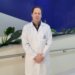Dr. Daniel Beltrame, urologista do Hospital de Caridade São Vicente de Paulo, posa para foto em corredor do hospital, usando jaleco branco diante de uma parede azul e cinza com plantas decorativas ao fundo.