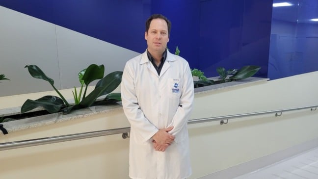 Dr. Daniel Beltrame, urologista do Hospital de Caridade São Vicente de Paulo, posa para foto em corredor do hospital, usando jaleco branco diante de uma parede azul e cinza com plantas decorativas ao fundo.