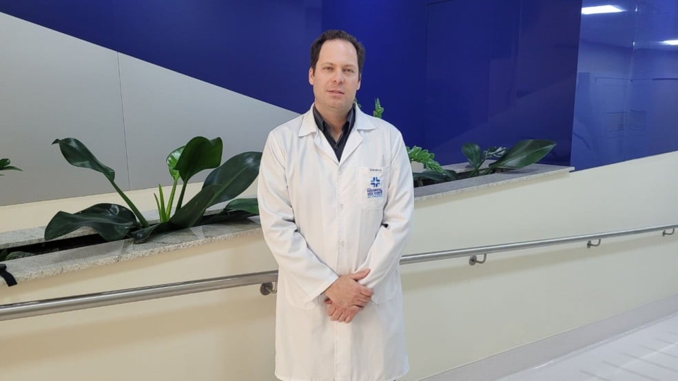 Dr. Daniel Beltrame, urologista do Hospital de Caridade São Vicente de Paulo, posa para foto em corredor do hospital, usando jaleco branco diante de uma parede azul e cinza com plantas decorativas ao fundo. Dr. Daniel Beltrame, urologista do Hospital de Caridade São Vicente de Paulo, posa para foto em corredor do hospital, usando jaleco branco diante de uma parede azul e cinza com plantas decorativas ao fundo.