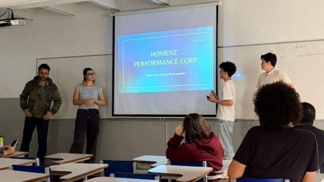 Grupo de estudantes da ESAMC Jundiaí apresenta um projeto diante da turma, com um slide projetado intitulado “Homenz Performance Corp”.