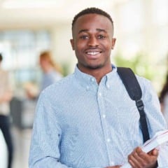 Estudante segurando cadernos e mochila em um corredor universitário, sorrindo para a câmera.