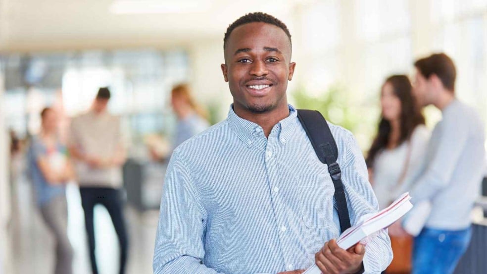 Estudante segurando cadernos e mochila em um corredor universitário, sorrindo para a câmera.