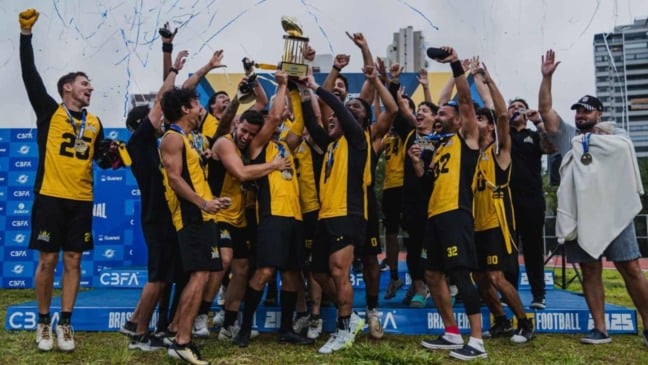 Time do Flag Kings comemora título nacional no palco, com jogadores levantando troféu e exibindo medalhas sob chuva de confetes.