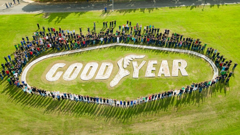 Vista aérea de colaboradores da Goodyear formando um círculo ao redor do logotipo da empresa no gramado, com a palavra “GOODYEAR” e o símbolo da asa alada. Vista aérea de colaboradores da Goodyear formando um círculo ao redor do logotipo da empresa no gramado, com a palavra “GOODYEAR” e o símbolo da asa alada.