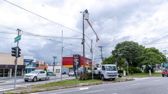 Trabalhador em cesto aéreo instala luminária de LED em poste alto em cruzamento movimentado de Jundiaí, com veículos e comércios ao fundo sob céu nublado.