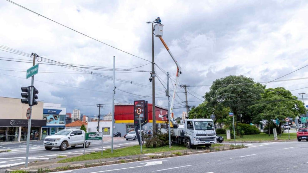 Trabalhador em cesto aéreo instala luminária de LED em poste alto em cruzamento movimentado de Jundiaí, com veículos e comércios ao fundo sob céu nublado. Trabalhador em cesto aéreo instala luminária de LED em poste alto em cruzamento movimentado de Jundiaí, com veículos e comércios ao fundo sob céu nublado.