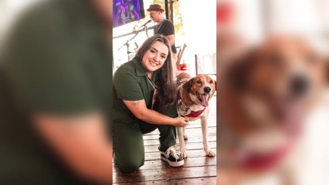 Mulher abaixada sorri ao lado de um cachorro de médio porte com lenço colorido, em um evento com música ao vivo ao fundo.