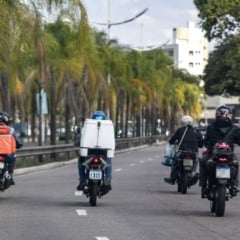 Entregadores de moto circulando por avenida arborizada em Jundiaí, com mochilas de delivery nas costas.