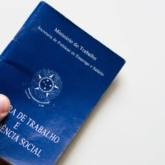 Mão segurando uma Carteira de Trabalho e Previdência Social azul, com o brasão do Brasil em destaque sobre fundo branco.