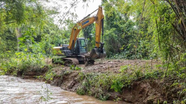 Escavadeira remove sedimentos nas margens de um rio cercado por vegetação, durante operação de desassoreamento em Jundiaí.