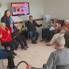 Idosos da Cidade Vicentina Frederico Ozanam participam de atividade recreativa em grupo com argolas coloridas, acompanhados por cuidadoras, em um ambiente acolhedor e iluminado.