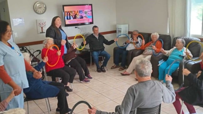 Idosos da Cidade Vicentina Frederico Ozanam participam de atividade recreativa em grupo com argolas coloridas, acompanhados por cuidadoras, em um ambiente acolhedor e iluminado.
