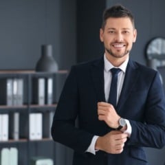Homem de terno sorrindo e ajustando o punho da camisa em escritório corporativo moderno.