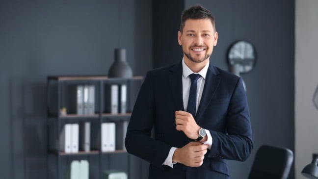 Homem de terno sorrindo e ajustando o punho da camisa em escritório corporativo moderno.