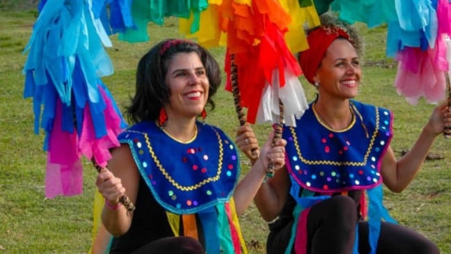 Aline Longui e Josiane Gomes, vestidas com trajes coloridos e segurando bastões decorados com fitas, participam de atividade do projeto Andanças do Ser-Tão Brincante em área verde de Jundiaí.