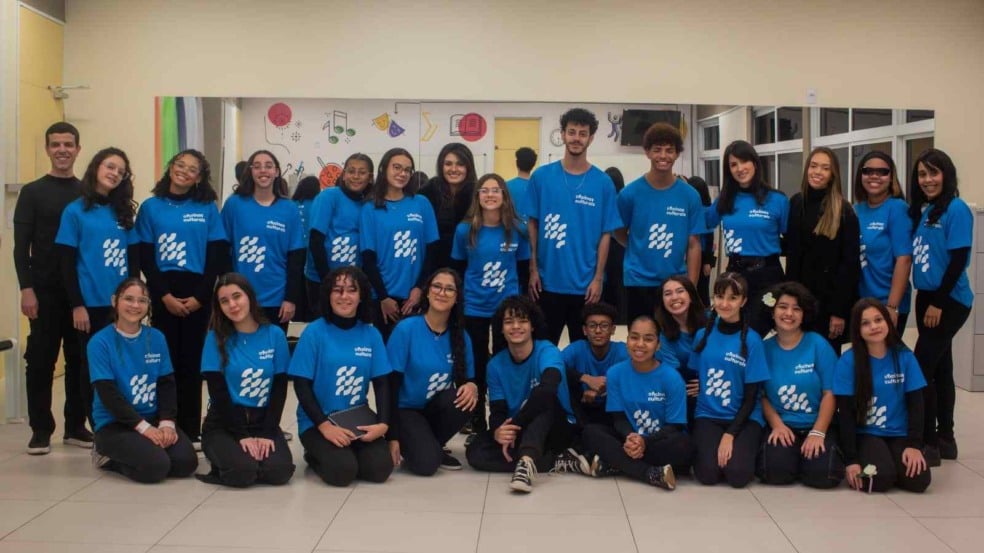 Coral juvenil das Oficinas Culturais reunido em sala de ensaio, com cerca de 25 jovens usando camisetas azuis e posando para foto em grupo. Coral juvenil das Oficinas Culturais reunido em sala de ensaio, com cerca de 25 jovens usando camisetas azuis e posando para foto em grupo.