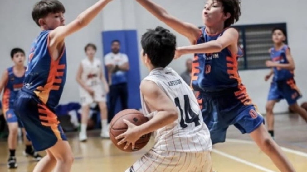 Primeira imagem: Menino com uniforme branco número 14 da RM Stars segura a bola em posição de ataque enquanto dois jogadores adversários de azul tentam bloqueá-lo durante uma partida de basquete.