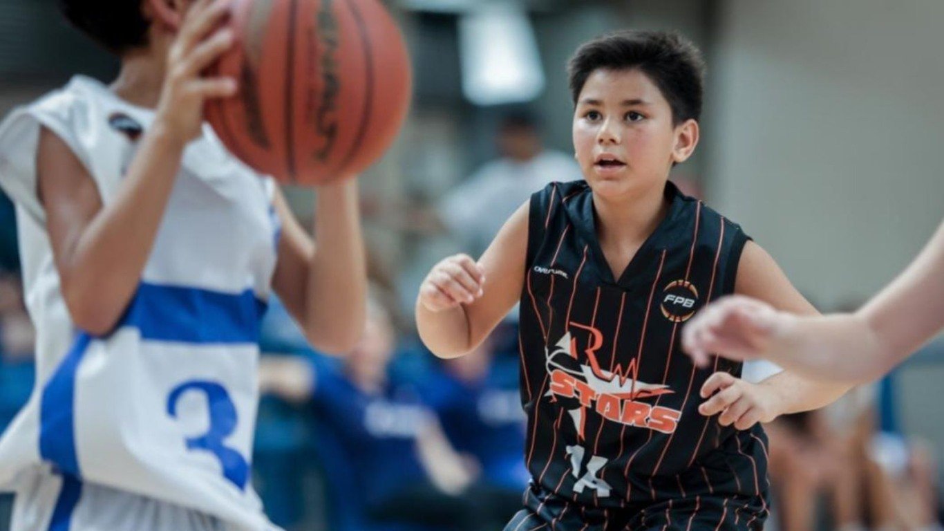 Terceira imagem: Jogador de basquete da RM Stars observa atentamente o adversário enquanto se posiciona na defesa, durante uma partida em quadra coberta.