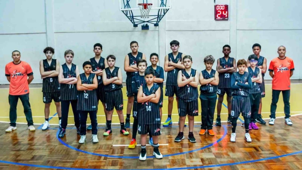 Segunda imagem: Equipe infantil da RM Stars posa para foto oficial de basquete em quadra coberta, com 16 jogadores uniformizados em preto e dois treinadores com camisetas laranja ao fundo.
