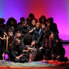 Grupo de atores vestidos de preto cercam um intérprete sentado no chão, em uma cena dramática do espetáculo “Até quando”, com iluminação colorida em tons de verde, vermelho e roxo ao fundo.