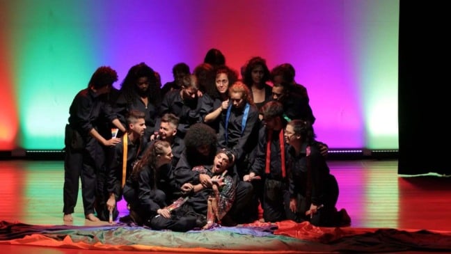 Grupo de atores vestidos de preto cercam um intérprete sentado no chão, em uma cena dramática do espetáculo “Até quando”, com iluminação colorida em tons de verde, vermelho e roxo ao fundo.