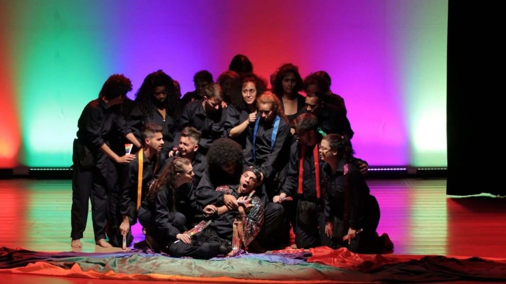 Grupo de atores vestidos de preto cercam um intérprete sentado no chão, em uma cena dramática do espetáculo “Até quando”, com iluminação colorida em tons de verde, vermelho e roxo ao fundo.