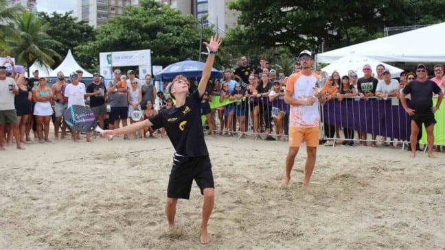Jovem atleta se prepara para sacar em uma partida de beach tennis na praia, observado por público atrás das grades e outro jogador aguardando a jogada.