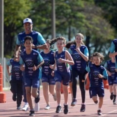 Crianças e adolescentes correm sorridentes em pista de atletismo, acompanhados por treinador, durante atividade esportiva ao ar livre em Jundiaí.