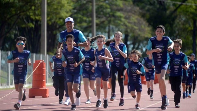 Crianças e adolescentes correm sorridentes em pista de atletismo, acompanhados por treinador, durante atividade esportiva ao ar livre em Jundiaí.