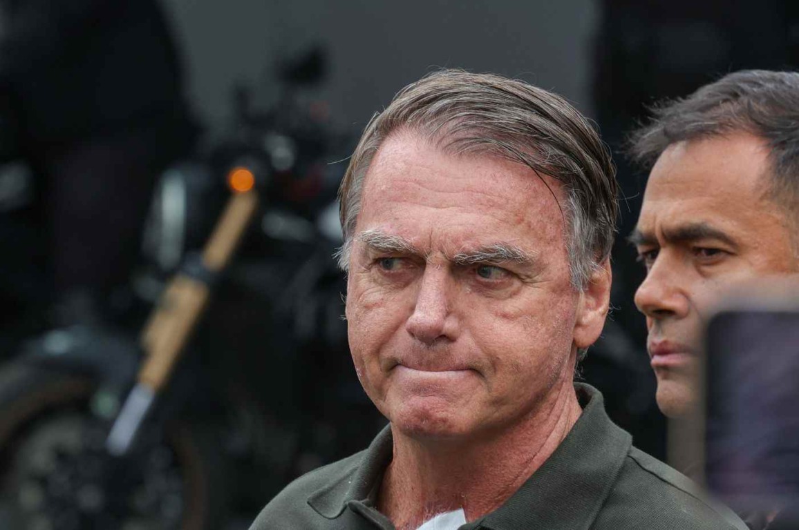 Ex-presidente Jair Bolsonaro de expressão séria, com a cabeça levemente inclinada para baixo, enquanto outra pessoa aparece parcialmente ao fundo.
