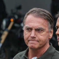 Ex-presidente Jair Bolsonaro de expressão séria, com a cabeça levemente inclinada para baixo, enquanto outra pessoa aparece parcialmente ao fundo.