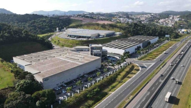 Imagem aérea da unidade da Brasilata em Jundiaí, com galpões industriais, áreas verdes e a rodovia à direita.