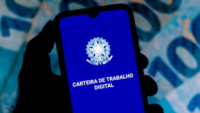 Celular mostrando o aplicativo Carteira de Trabalho Digital com o brasão do Brasil, em frente a notas de dinheiro desfocadas ao fundo.