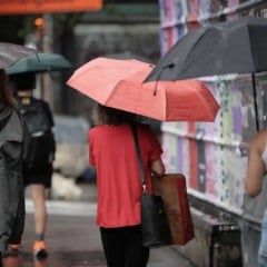 Pessoas com guarda-chuvas caminham em uma rua molhada durante chuva em área urbana.