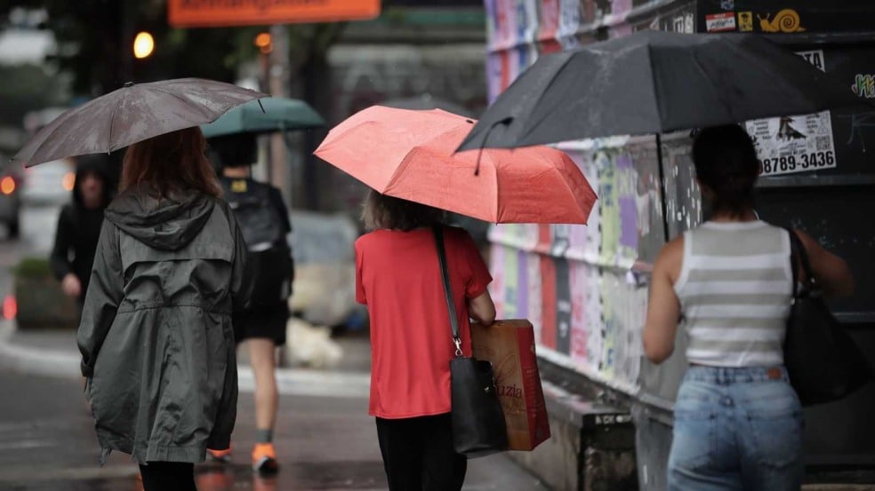 Pessoas com guarda-chuvas caminham em uma rua molhada durante chuva em área urbana.