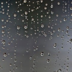 Gotas de chuva em um vidro com fundo cinza desfocado, simbolizando tempo nublado e possibilidade de chuva.
