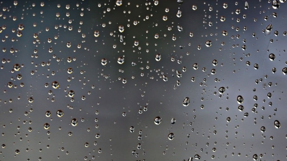 Gotas de chuva em um vidro com fundo cinza desfocado, simbolizando tempo nublado e possibilidade de chuva.