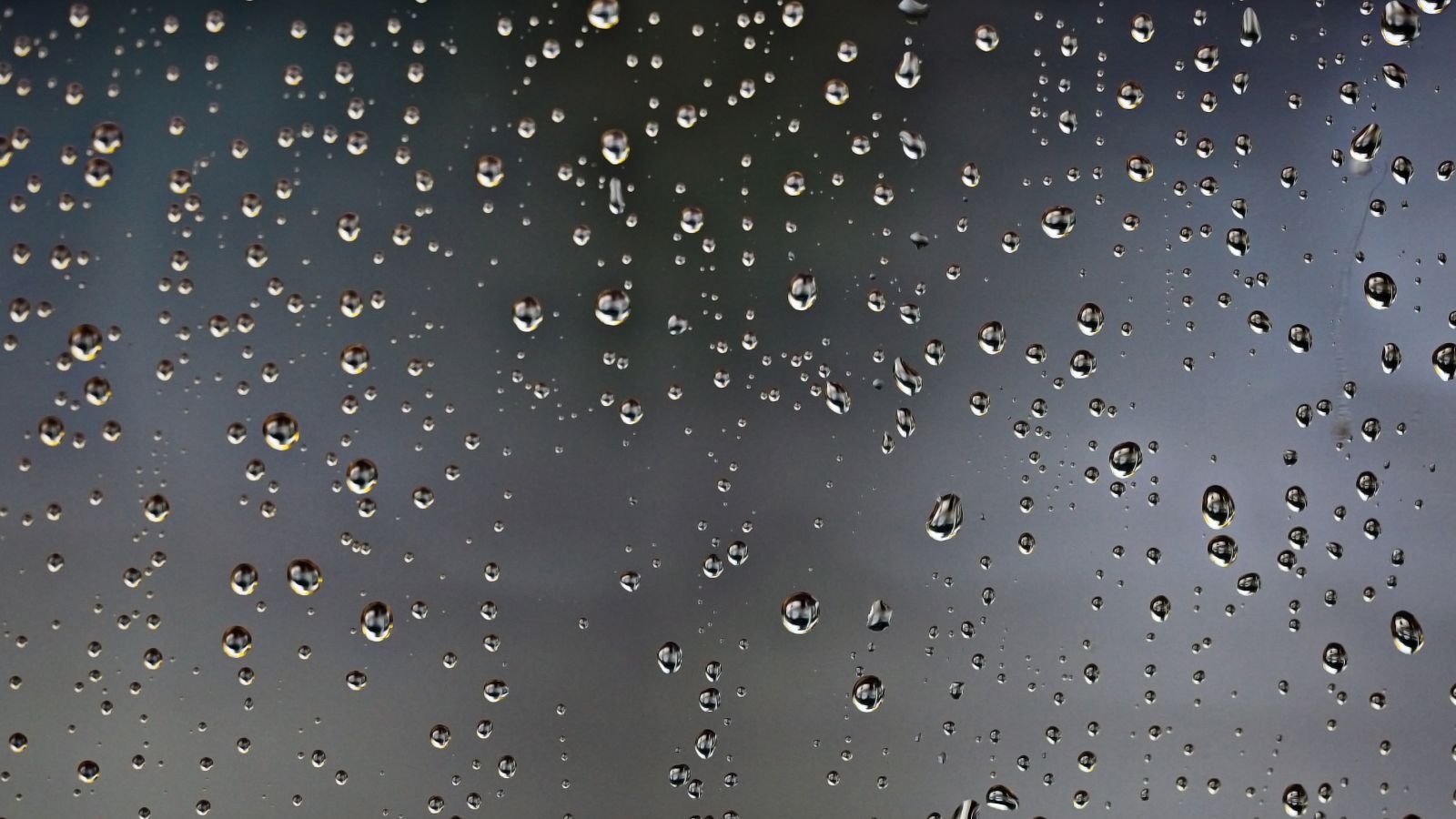 Gotas de chuva em um vidro com fundo cinza desfocado, simbolizando tempo nublado e possibilidade de chuva.