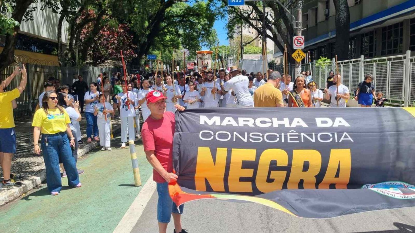 Faixa da Marcha da Consciência Negra sendo carregada por participantes, com grupo de capoeira tocando ao fundo e moradores acompanhando pelas calçadas.