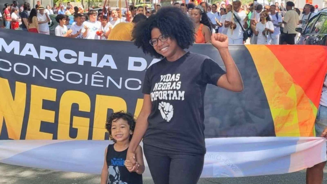Mulher com camiseta ‘Vidas Negras Importam’ marcha com uma criança diante da faixa da Marcha da Consciência Negra, levantando o punho em gesto de resistência.