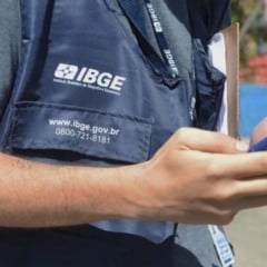Agente do IBGE de colete azul segura um celular durante coleta de dados em área externa.