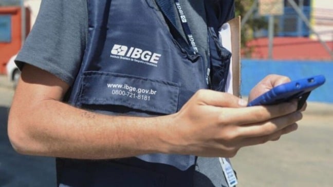 Agente do IBGE de colete azul segura um celular durante coleta de dados em área externa.