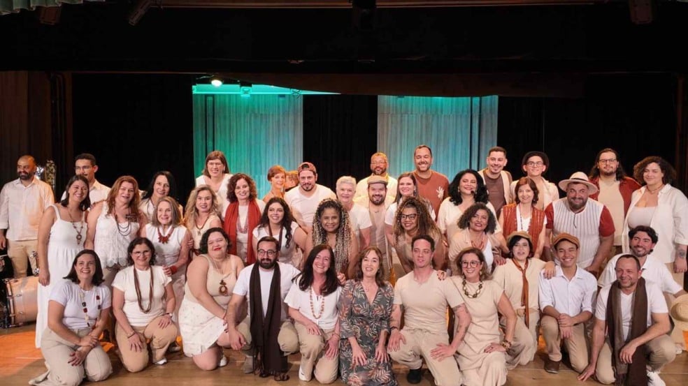 Foto de grupo com os integrantes do Coral Municipal de Jundiaí reunidos no palco, vestidos em tons claros e sorrindo, posados em várias fileiras diante de um fundo iluminado em verde. Foto de grupo com os integrantes do Coral Municipal de Jundiaí reunidos no palco, vestidos em tons claros e sorrindo, posados em várias fileiras diante de um fundo iluminado em verde.