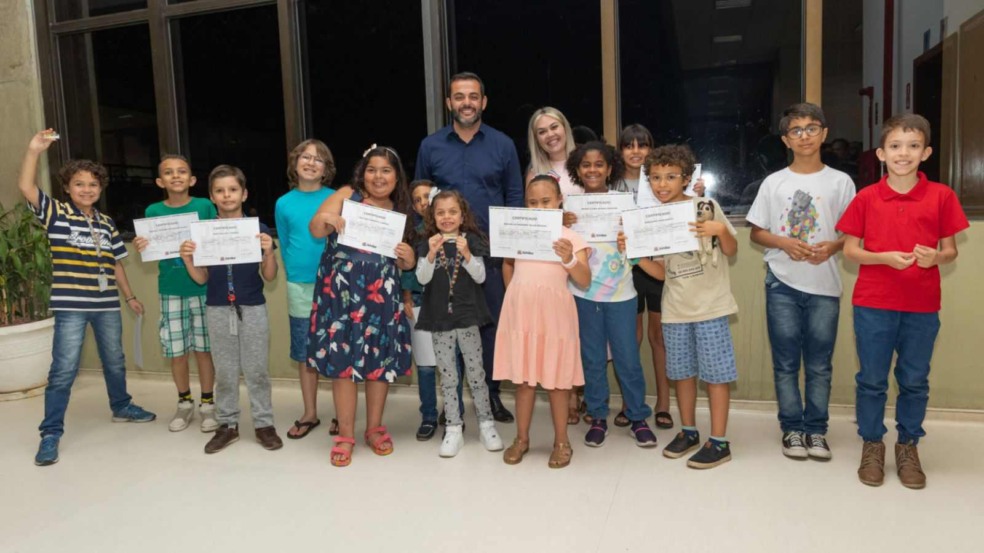 Crianças posam sorrindo com certificados e medalhas ao lado de um homem e uma mulher, em um ambiente interno com janelas ao fundo. Crianças posam sorrindo com certificados e medalhas ao lado de um homem e uma mulher, em um ambiente interno com janelas ao fundo.