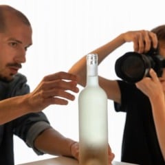 Pessoa ajustando uma garrafa enquanto outra fotografa o objeto com uma câmera profissional em estúdio.