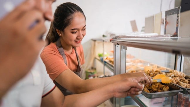 Mulher manipulando alimentos com pinça em vitrine de buffet, utilizando avental e seguindo boas práticas de higiene.