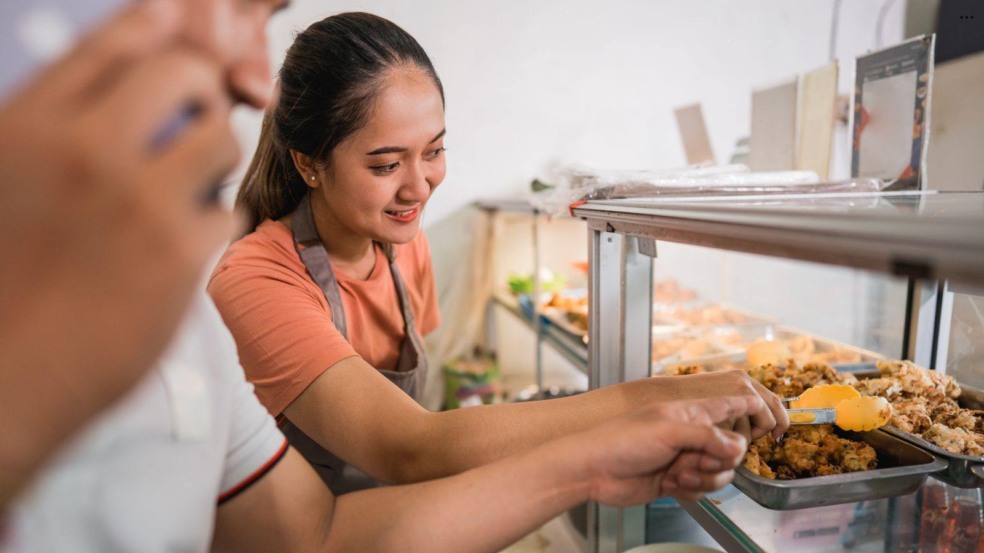 Mulher manipulando alimentos com pinça em vitrine de buffet, utilizando avental e seguindo boas práticas de higiene. Mulher manipulando alimentos com pinça em vitrine de buffet, utilizando avental e seguindo boas práticas de higiene.