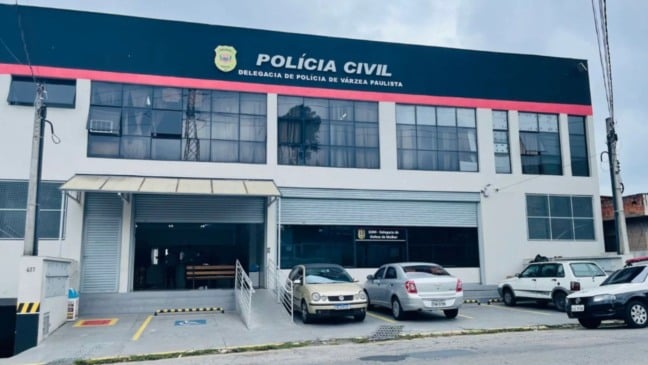 Fachada da Delegacia de Polícia de Várzea Paulista, com carros estacionados em frente e o letreiro da Polícia Civil visível no topo do prédio.